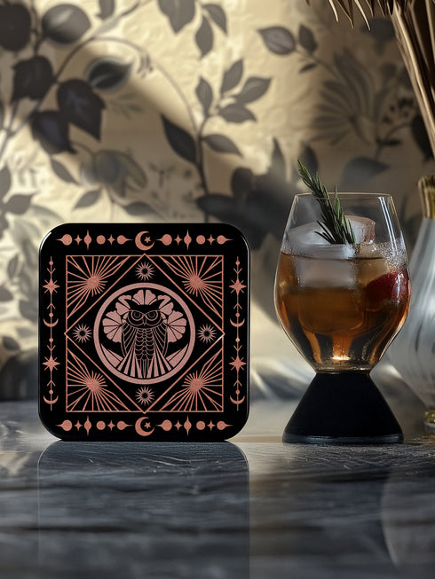A set of four black, high-gloss square coasters featuring intricate rose-gold metallic illustrations of an owl, raven, stag, and wolf framed by celestial and botanical motifs. Made from durable masonite hardboard with cork backing for slip-resistant protection.
